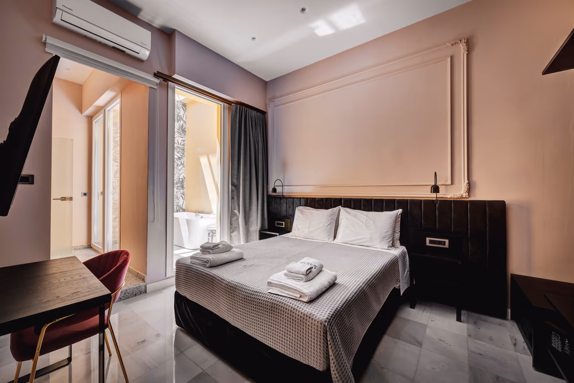 Modern hotel room with a double bed, white linens, folded towels, a red chair by a wooden desk, and an air conditioner above the window leading to a bathroom with a bathtub.