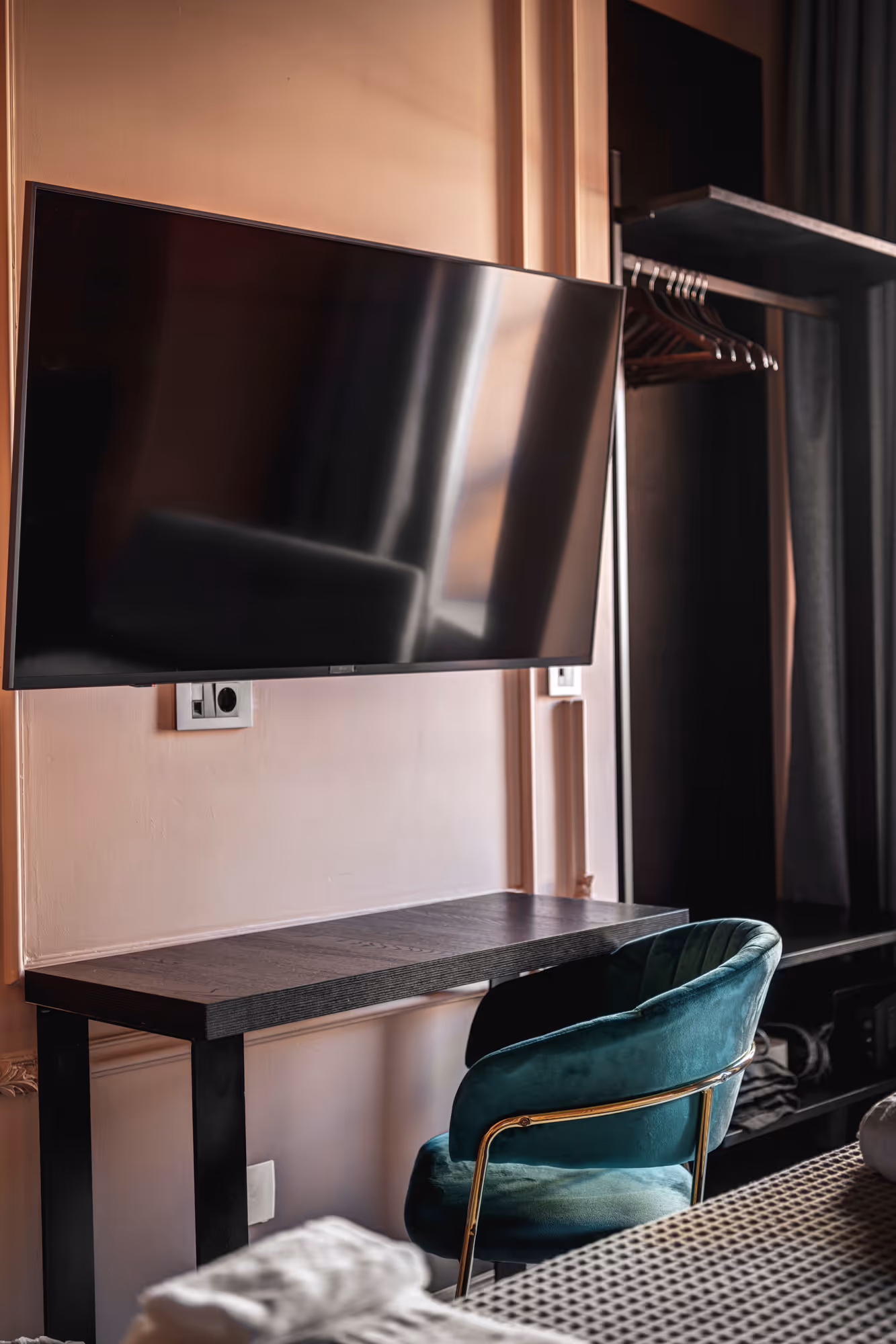 Modern hotel room corner with a wall-mounted flat-screen TV, dark wooden desk, and teal velvet chair with gold frame.