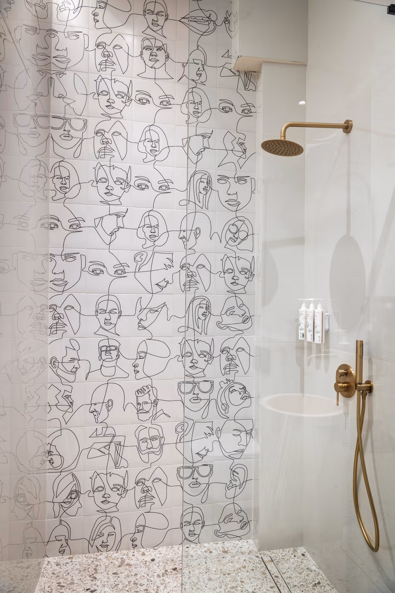 Shower area with gold fixtures and wall tiles featuring continuous line art faces in black on white background.