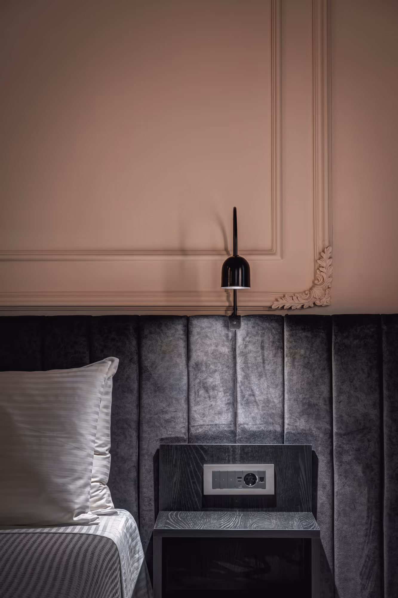 Bedside area with a black padded headboard, a white pillow on a striped bed, a black nightstand with electrical outlets, and a black wall-mounted lamp.