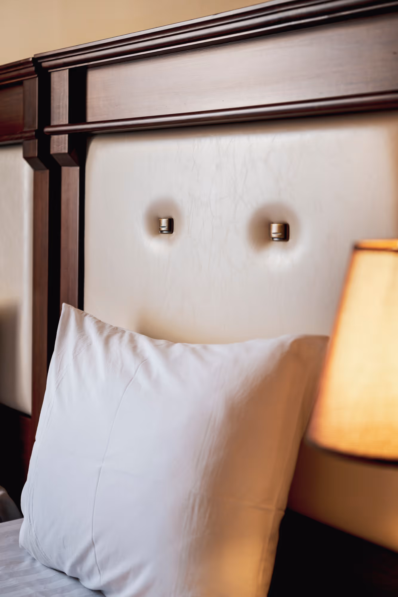 Close-up of a white pillow resting against a tufted cream-colored headboard with dark wood trim and a softly glowing lampshade on the right.