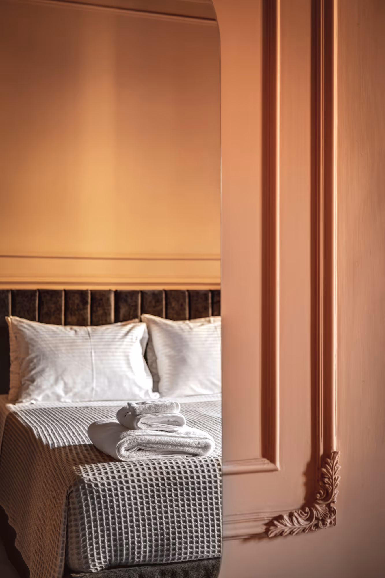Stack of white towels placed on a neatly made bed with white pillows and textured bedspread, seen through an open door with ornate molding.