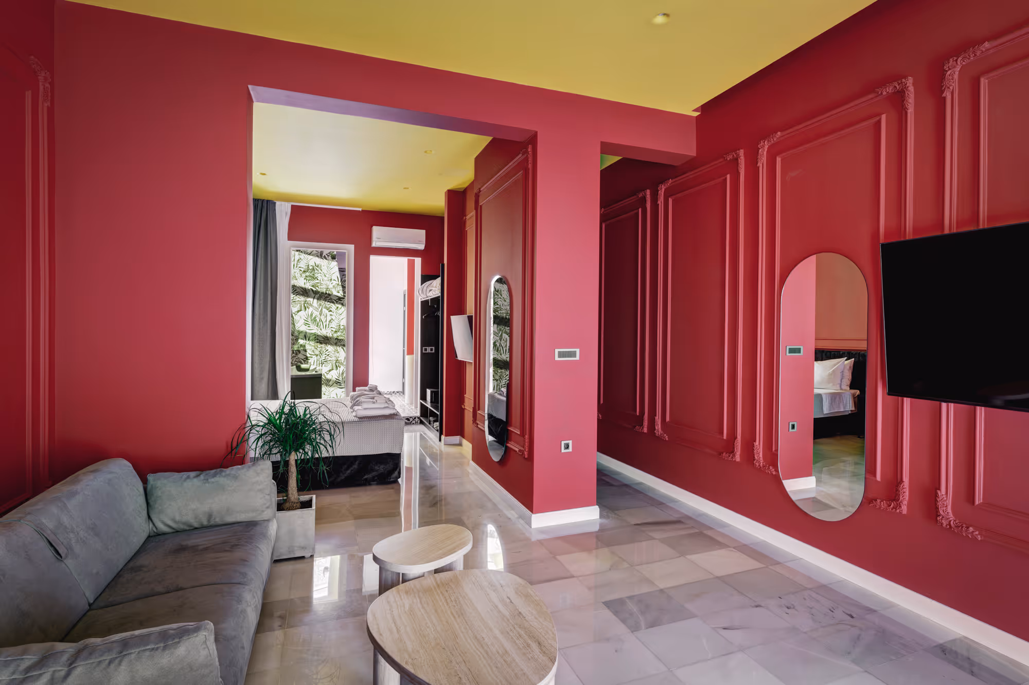 Modern living space with dark pink walls, yellow ceiling, gray sofa, two wooden coffee tables, a plant, wall-mounted TV, and a bedroom visible through an open doorway.