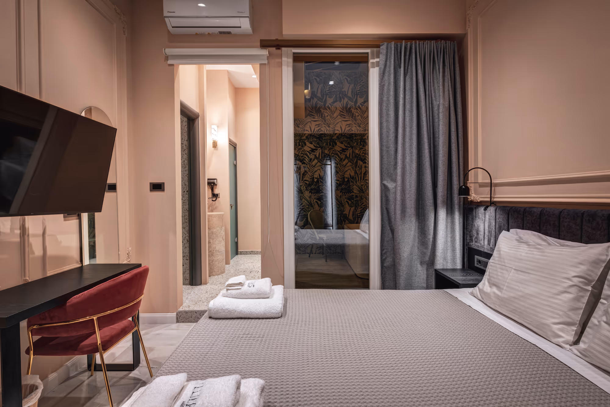 Hotel room with a double bed, gray bedding, white towels, a maroon chair, wall-mounted TV, and a visible bathroom entrance.