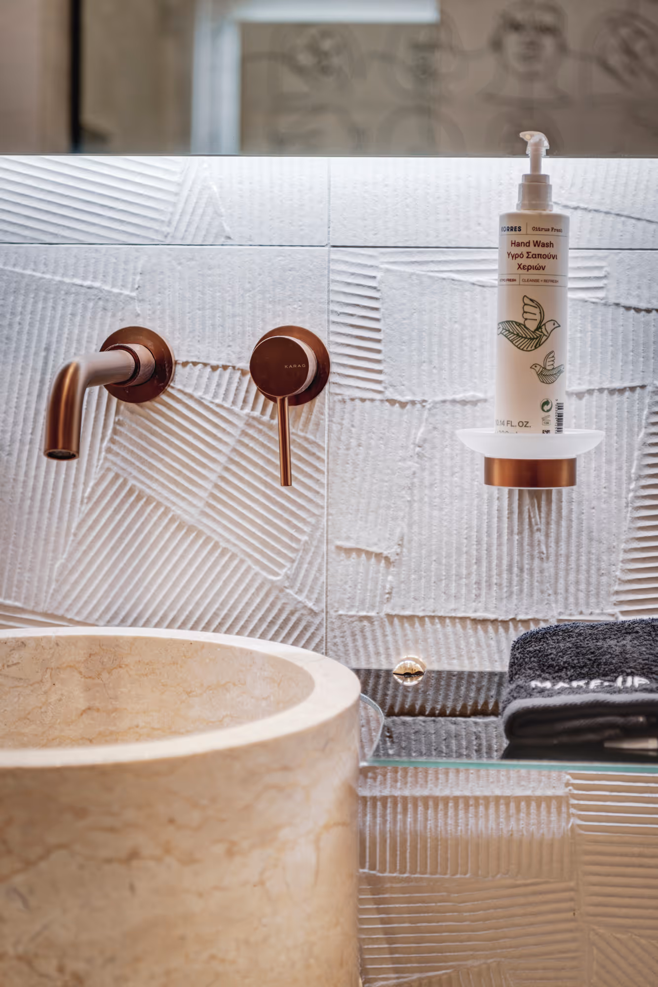 Modern bathroom sink with beige stone basin, bronze wall-mounted faucet, and hand wash dispenser on a glass shelf.