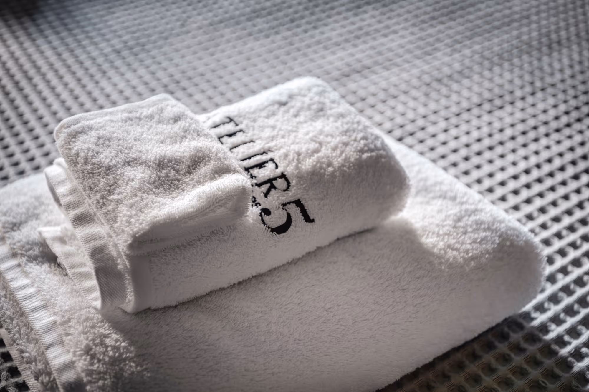 Stack of three white towels with one embroidered with 'TUIR5' in black thread on a textured surface.