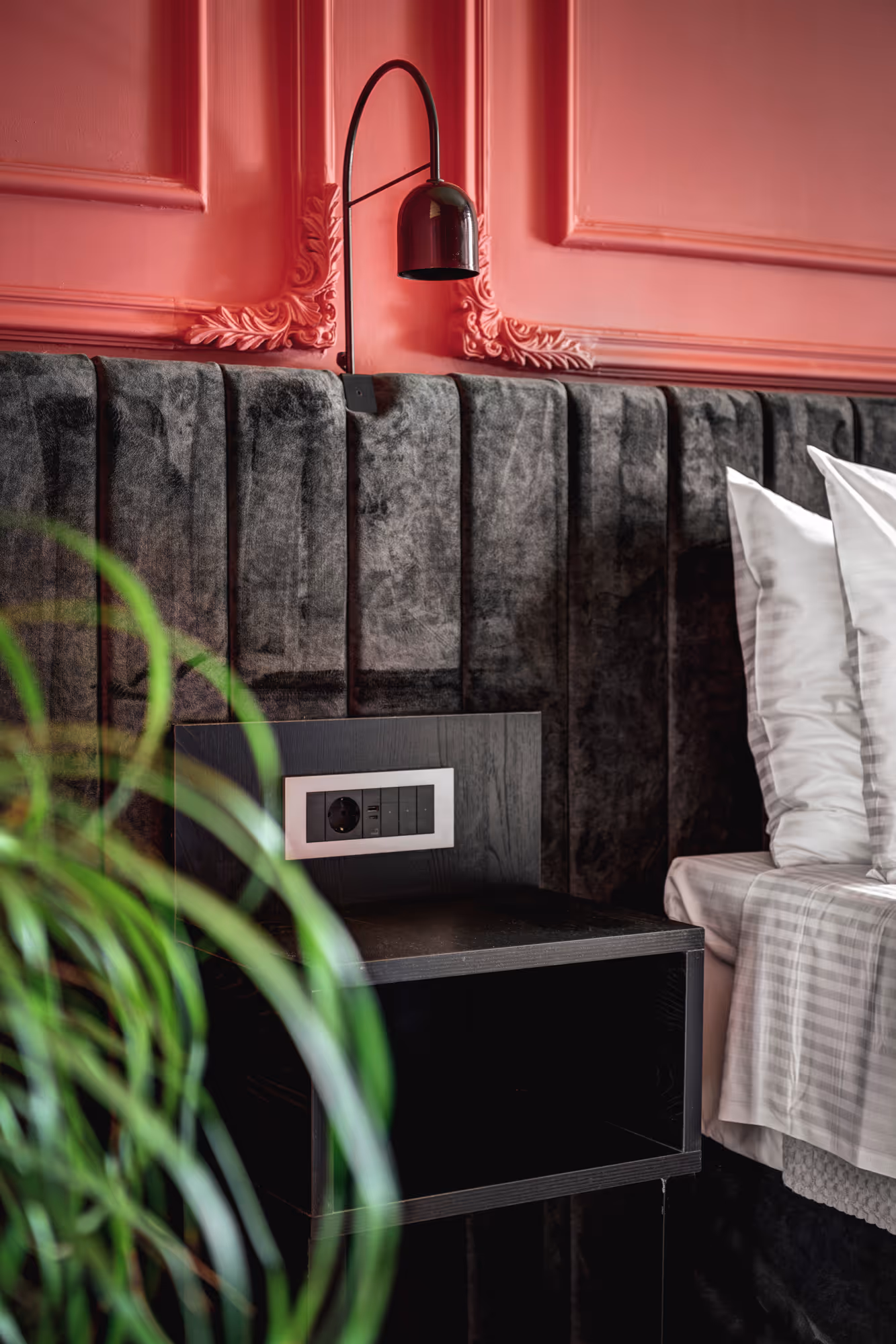 Close-up of a modern bedroom corner with a dark velvet padded headboard, pink wall with decorative molding, a black wall lamp, and a bedside table next to a bed with white pillows and gray striped bedding, with blurred green plant in the foreground.