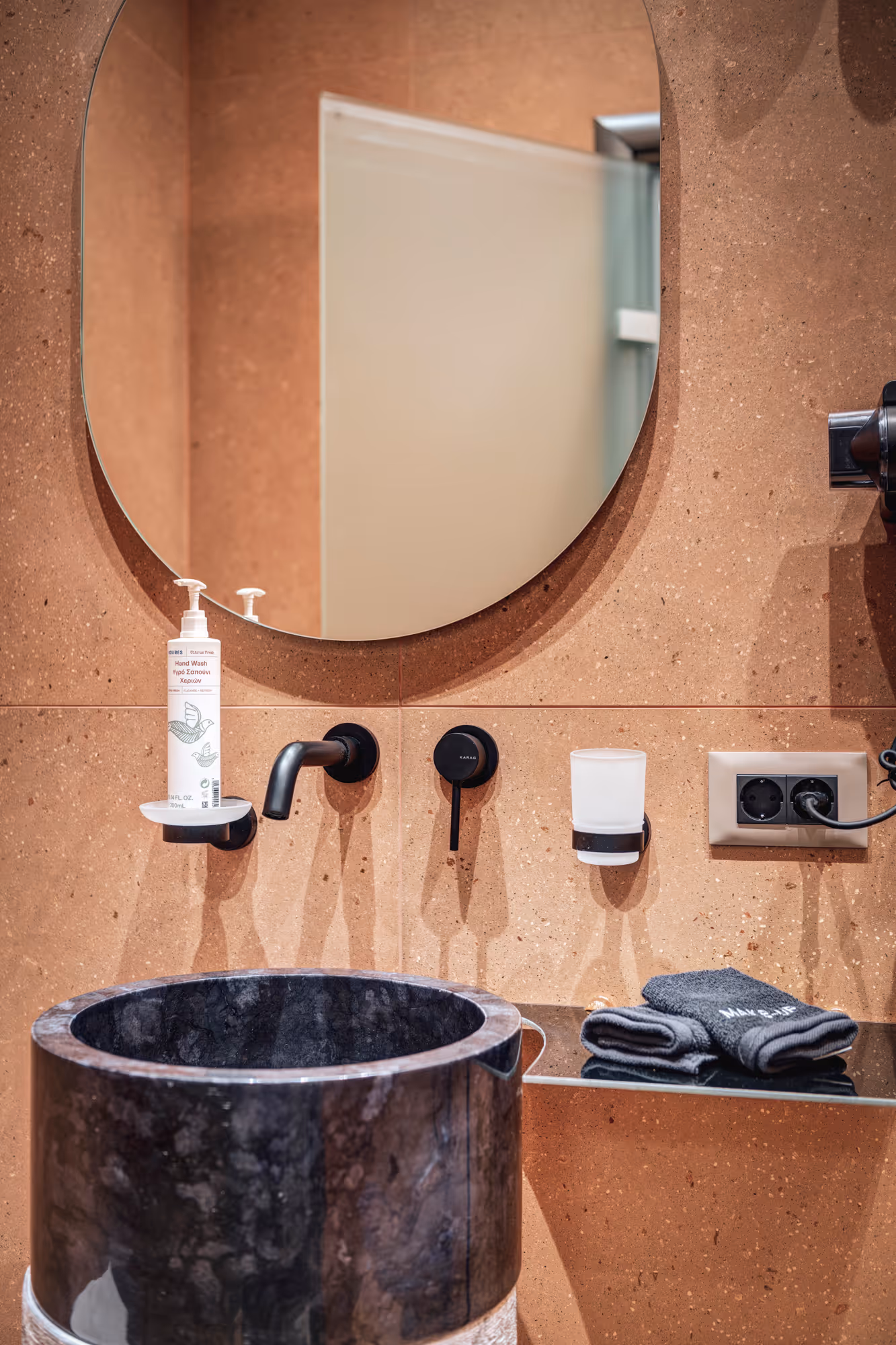 Modern bathroom sink with a round black marble basin, wall-mounted faucet and hand wash bottle, beige tiled wall, round mirror, and folded towels on a glass shelf.