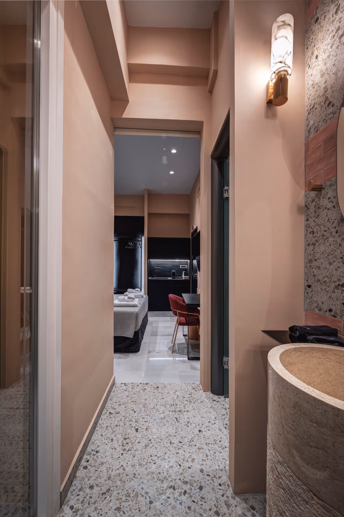 View down a hallway with terrazzo flooring leading to a bedroom with a bed, a red chair, and a kitchenette, and a bathroom sink area with a round stone basin and wall light on the right.