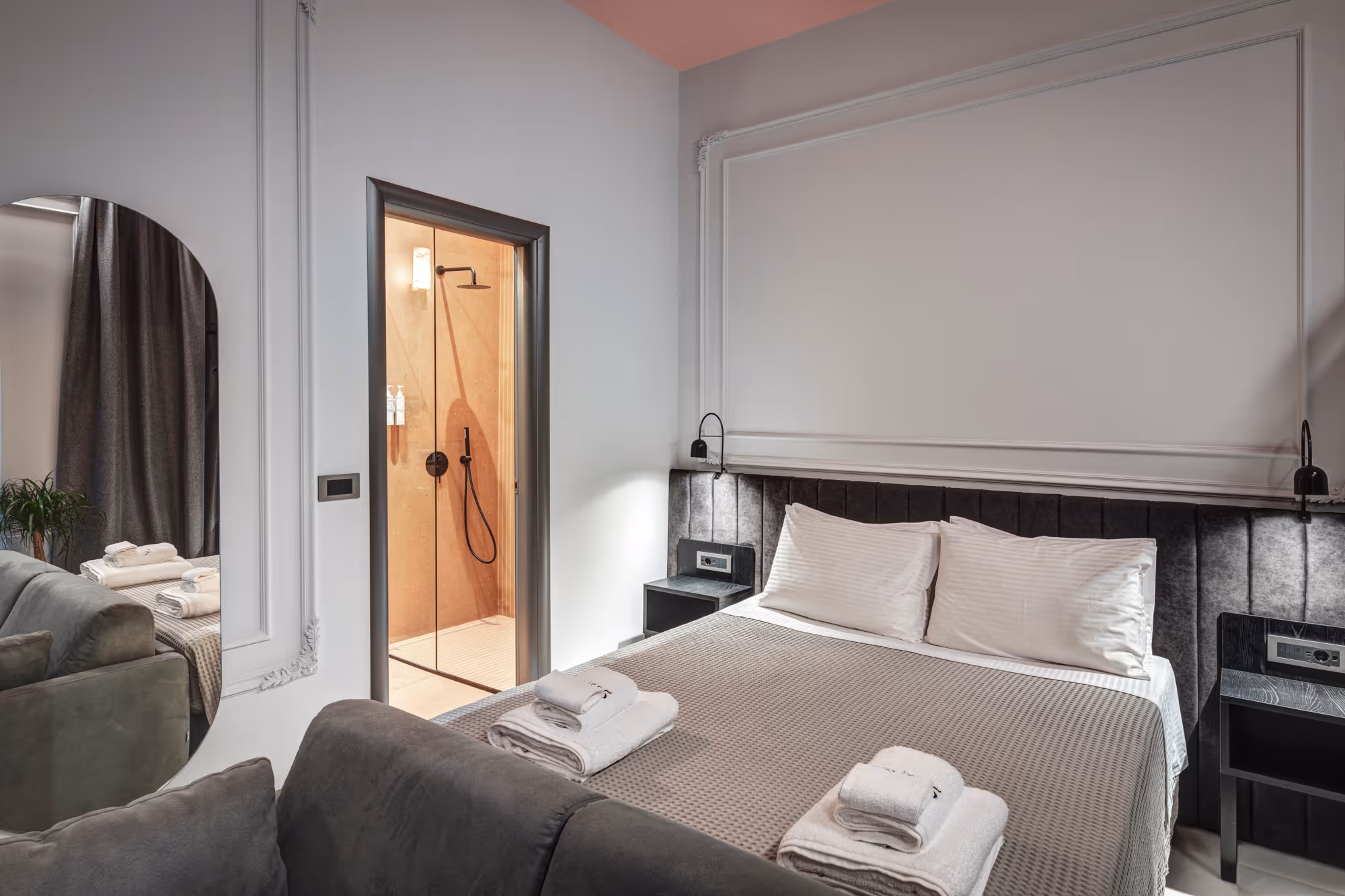 Modern bedroom with a large bed, gray upholstered headboard, folded white towels, and an open door leading to a shower area.