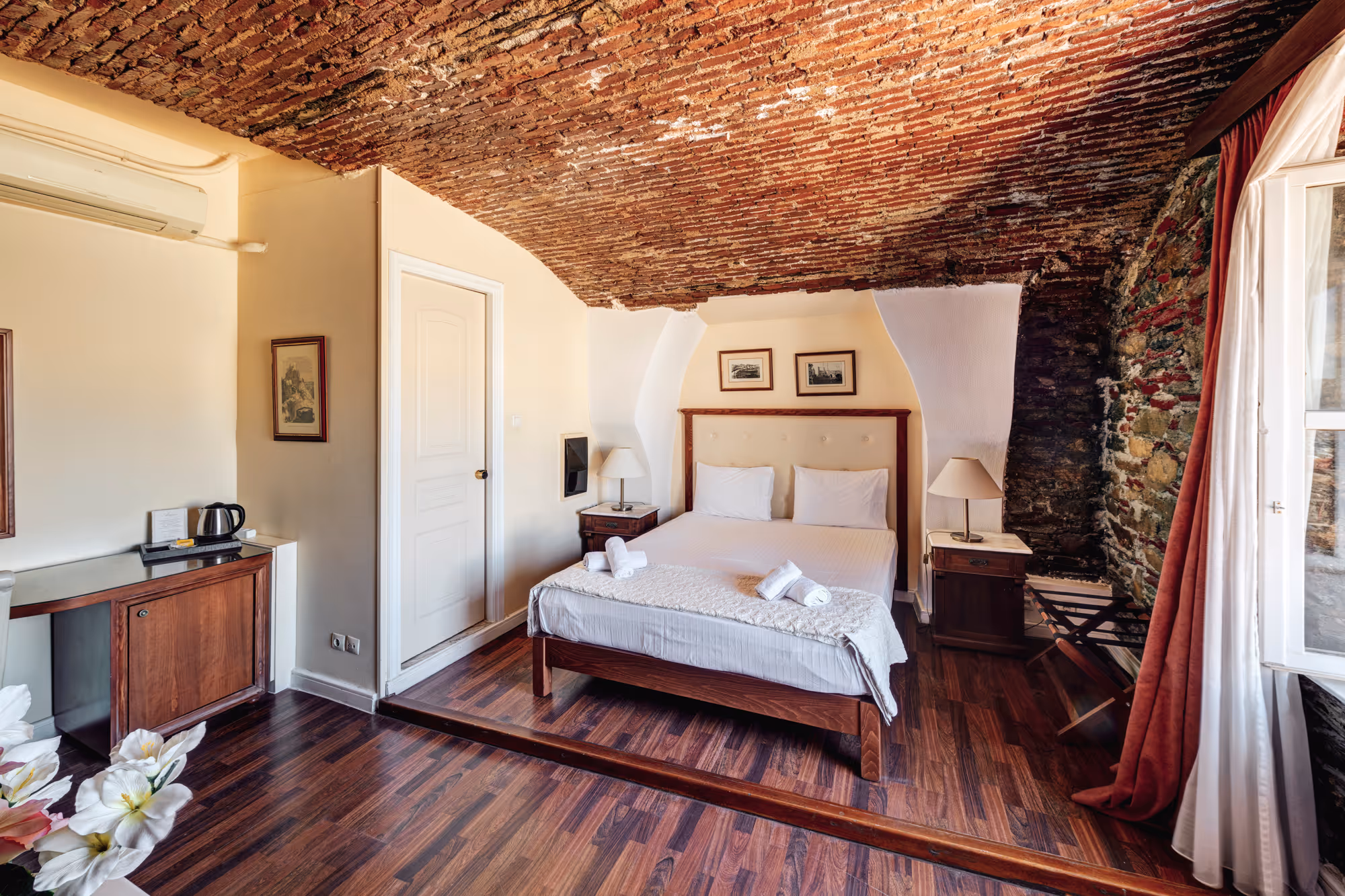 Cozy bedroom with exposed brick arched ceiling, wooden floor, double bed with white linens, and bedside tables with lamps.