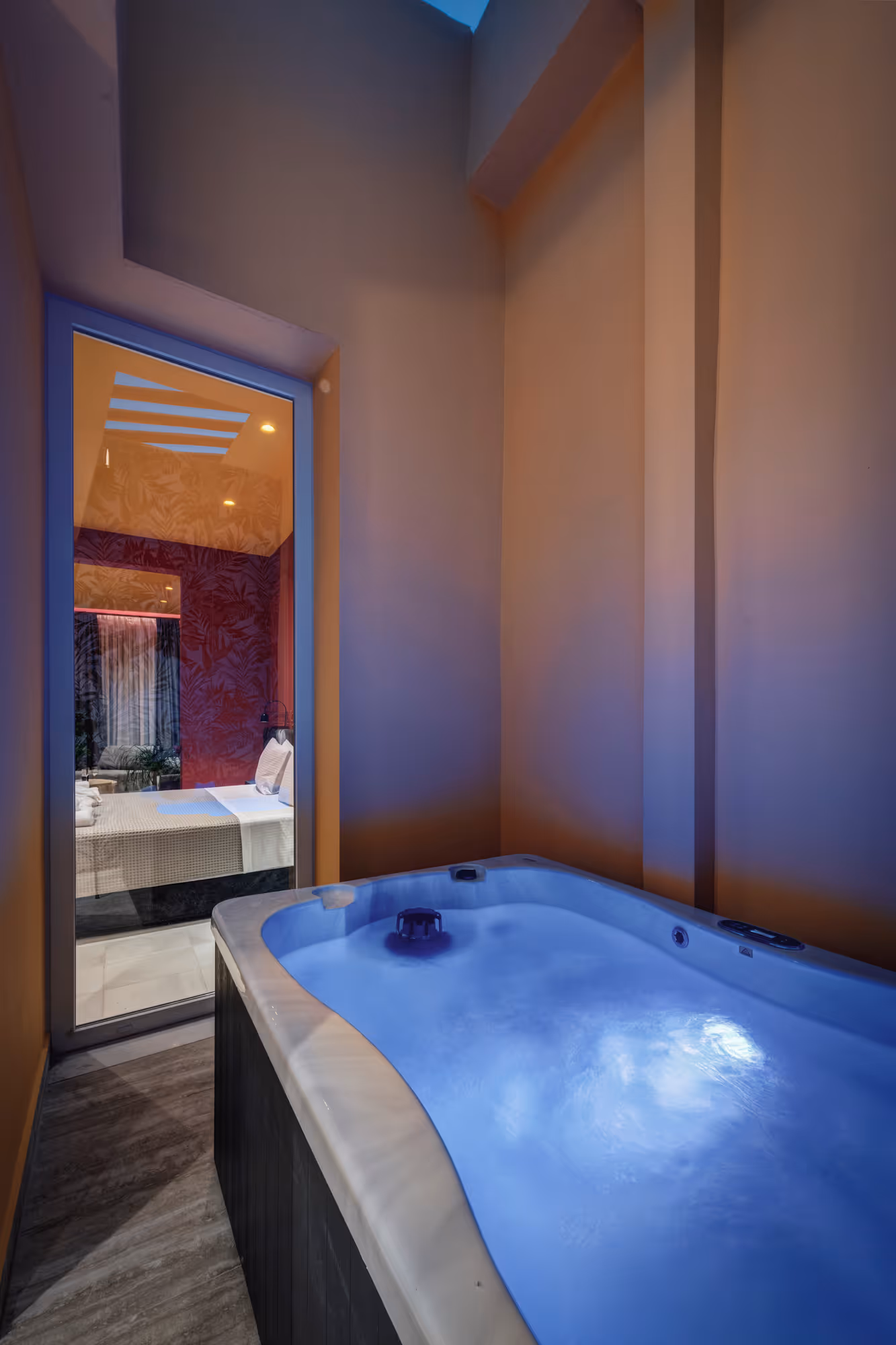 Indoor jacuzzi tub filled with blue-lit water next to a glass door showing a bedroom with a bed and patterned wallpaper.