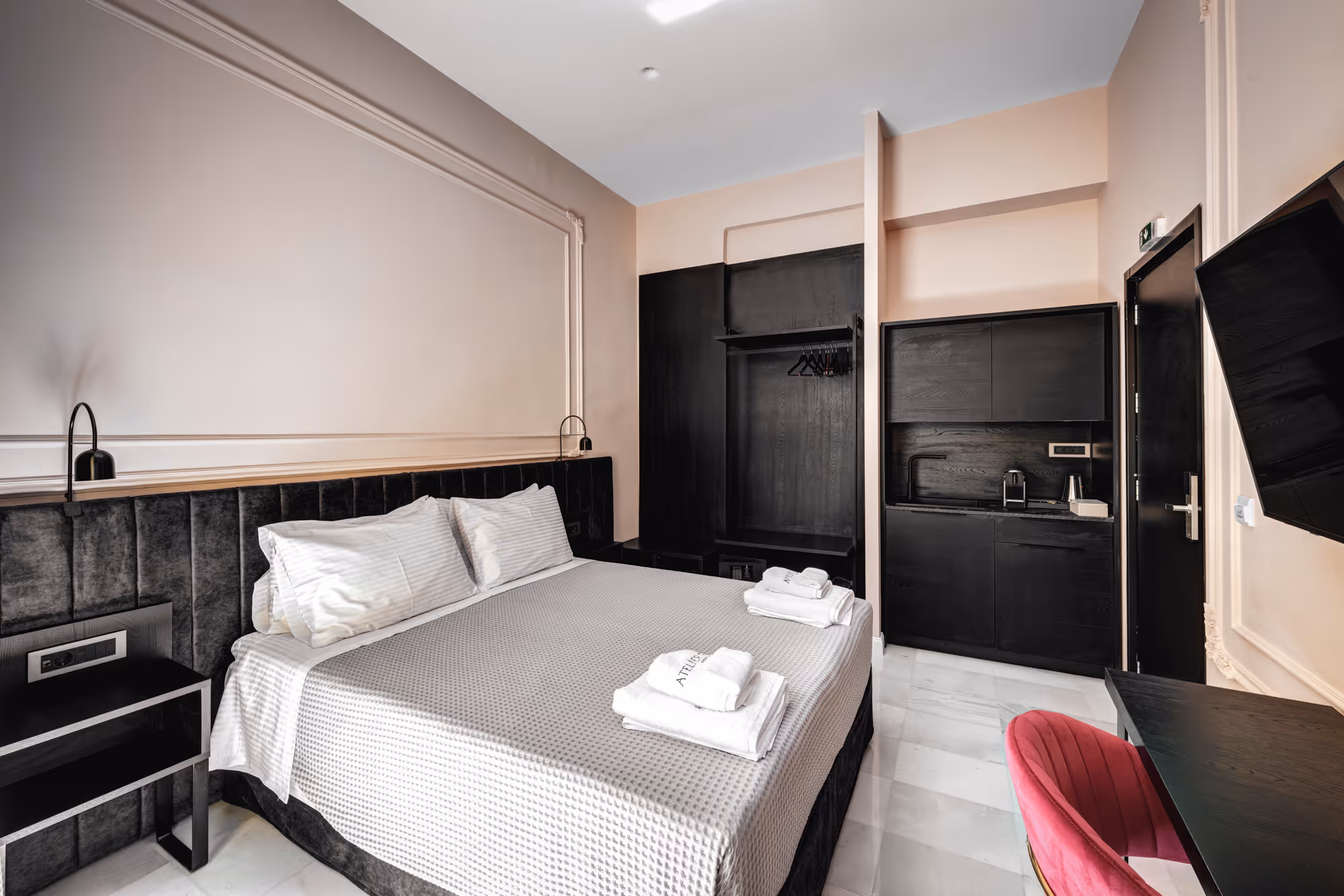 Modern hotel room with a large bed featuring white and gray bedding, black furniture, a small kitchenette, and a pink chair.