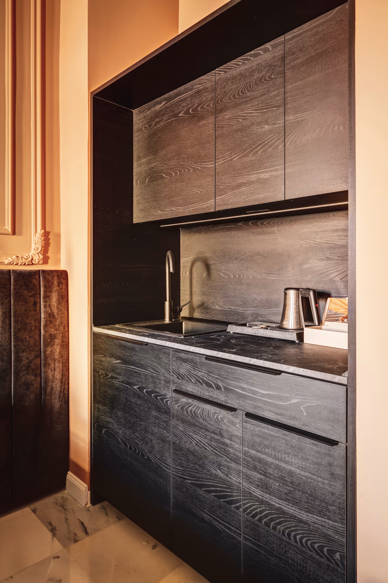 Compact modern kitchen with dark wood cabinets, black countertop, sink, and electric kettle on the right side.