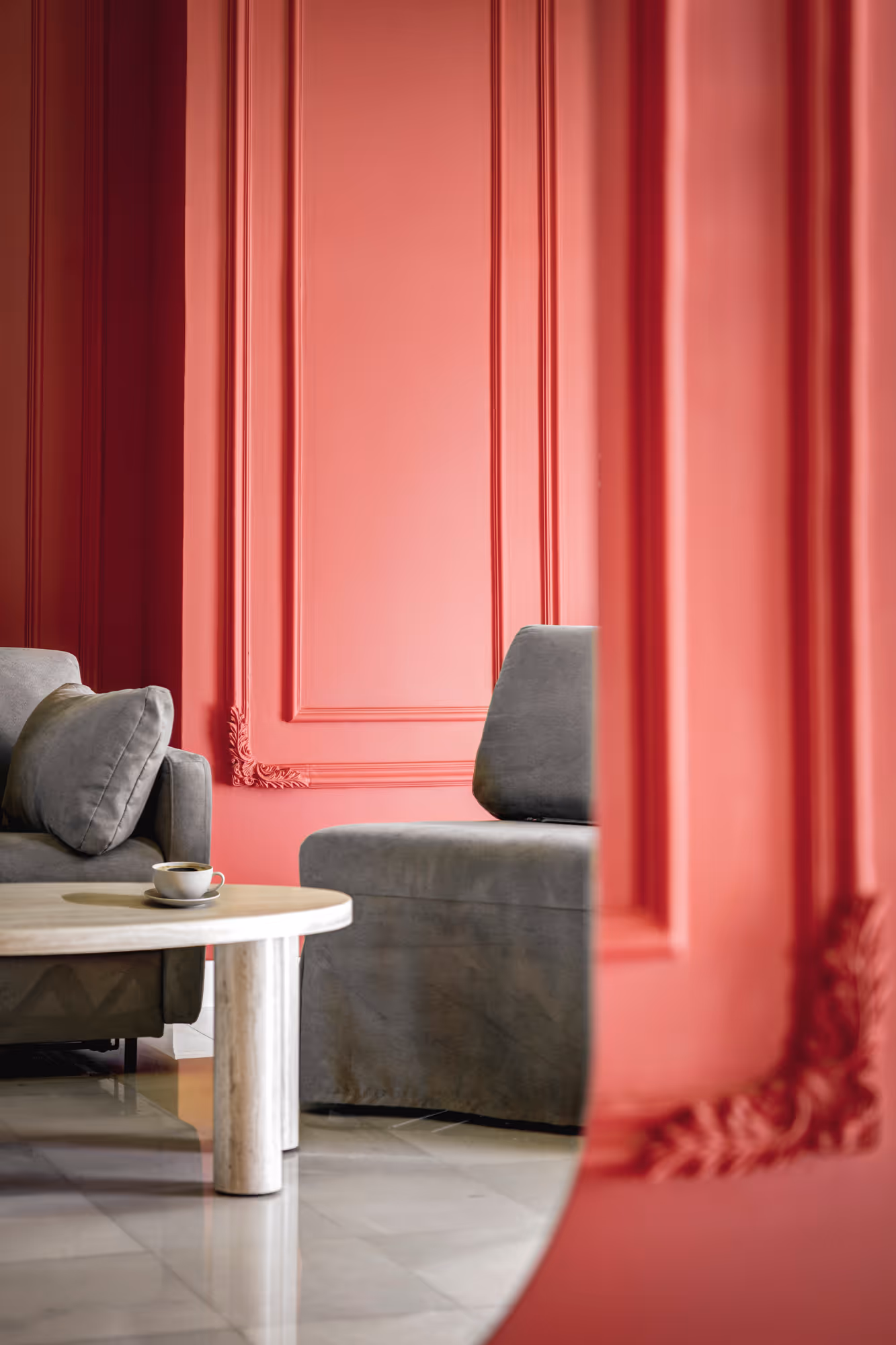 Modern living room corner with gray upholstered furniture, a round coffee table with a cup, and coral-colored walls with decorative molding.