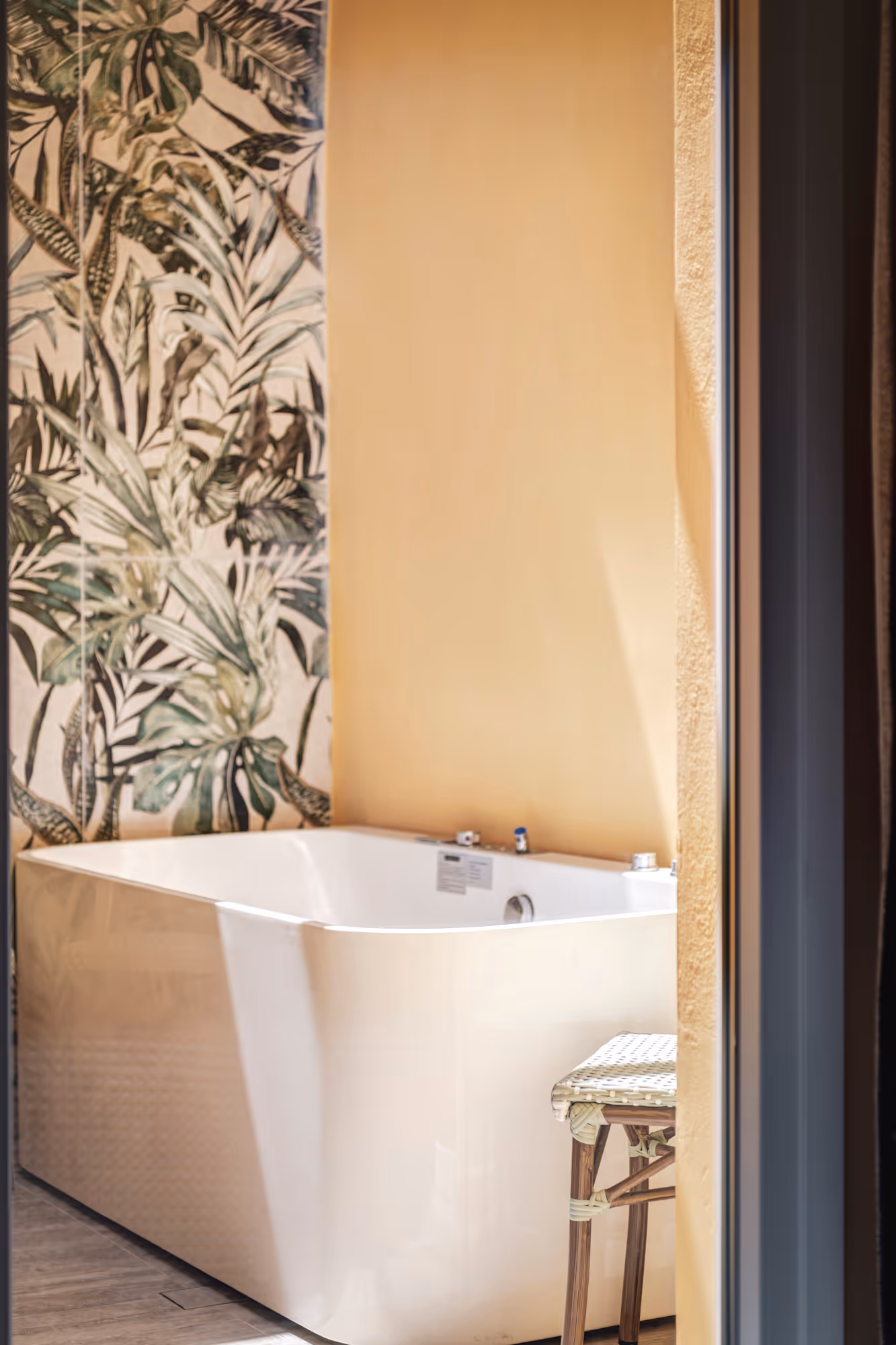 Modern white freestanding bathtub beside a tropical leaf patterned wall and a small wicker stool on wooden floor.