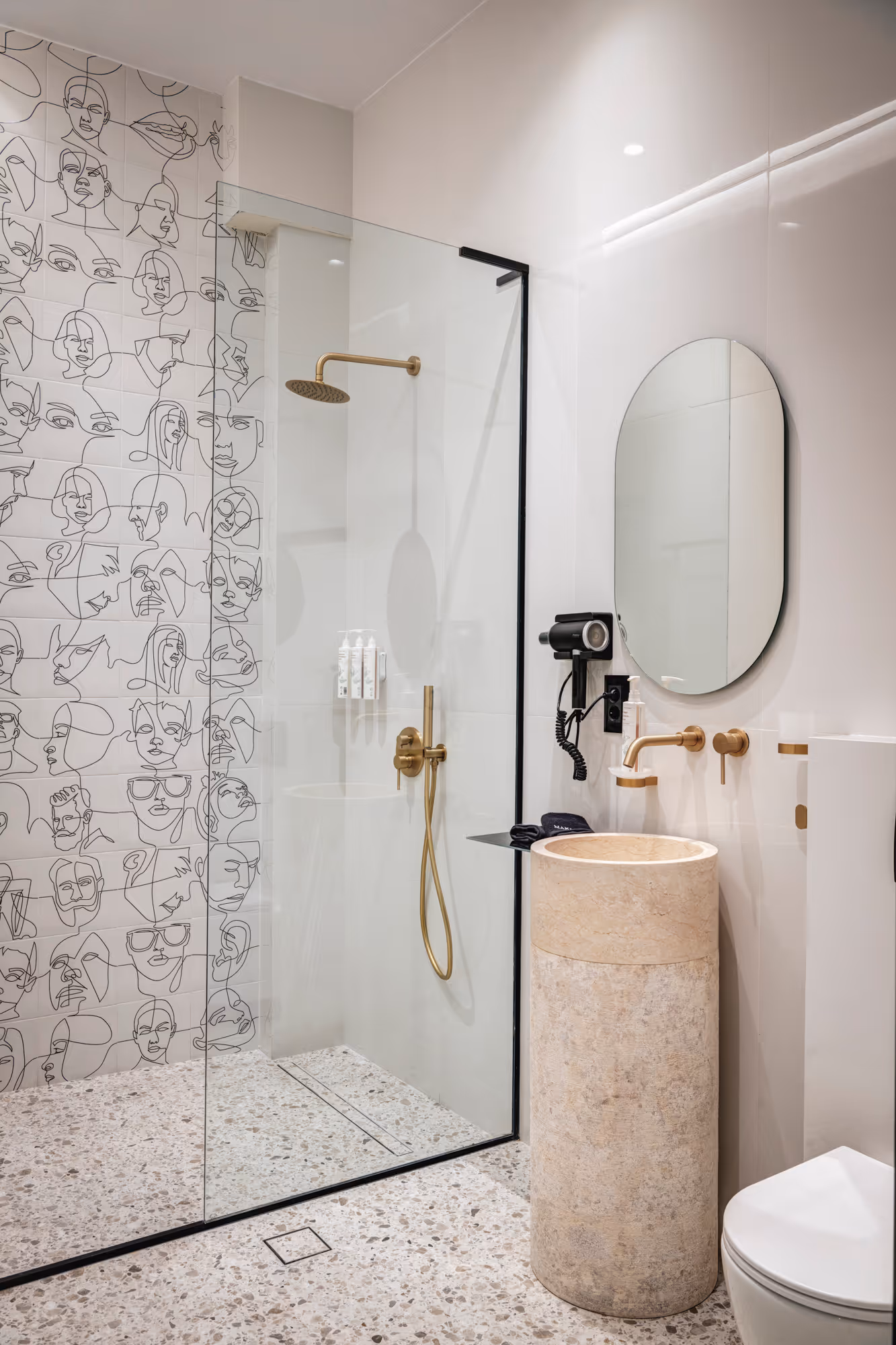 Modern bathroom with terrazzo floor, a walk-in glass shower with gold fixtures, line art face tiles, cylindrical stone sink, oval mirror, and wall-mounted hairdryer.