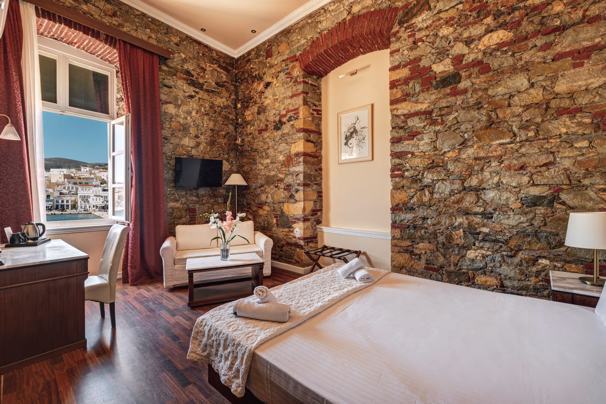 Cozy hotel room with stone walls, a large bed with white linens, a small white couch, wooden floor, and an open window showing a sea view and buildings.