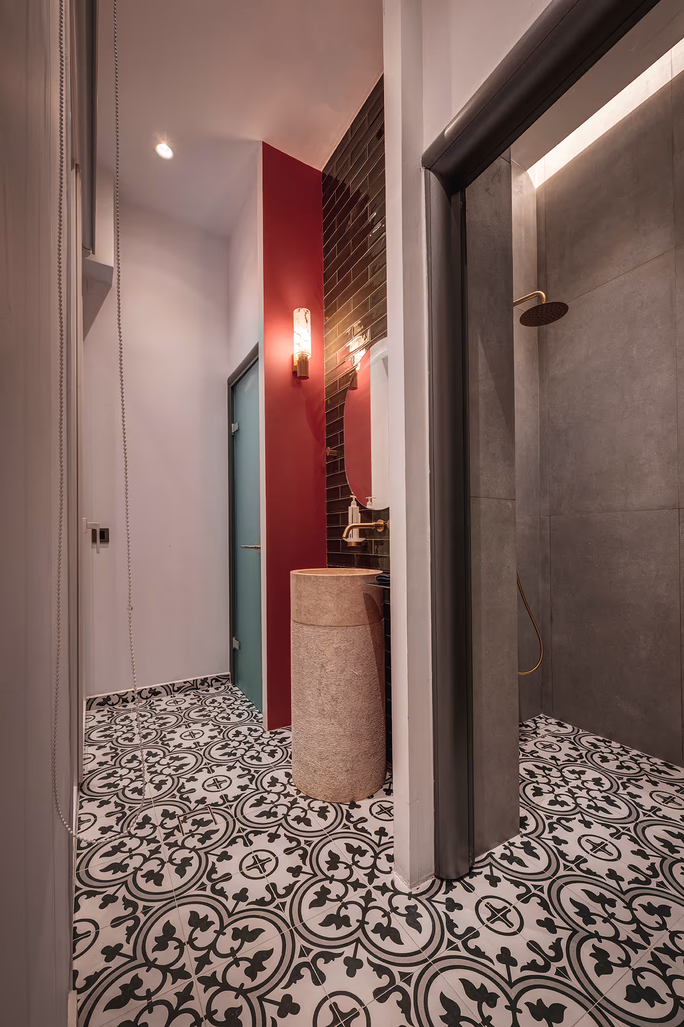 Modern bathroom with patterned black and white tiled floor, cylindrical stone sink, dark wall tiles, red accent wall, and walk-in shower with brass showerhead.