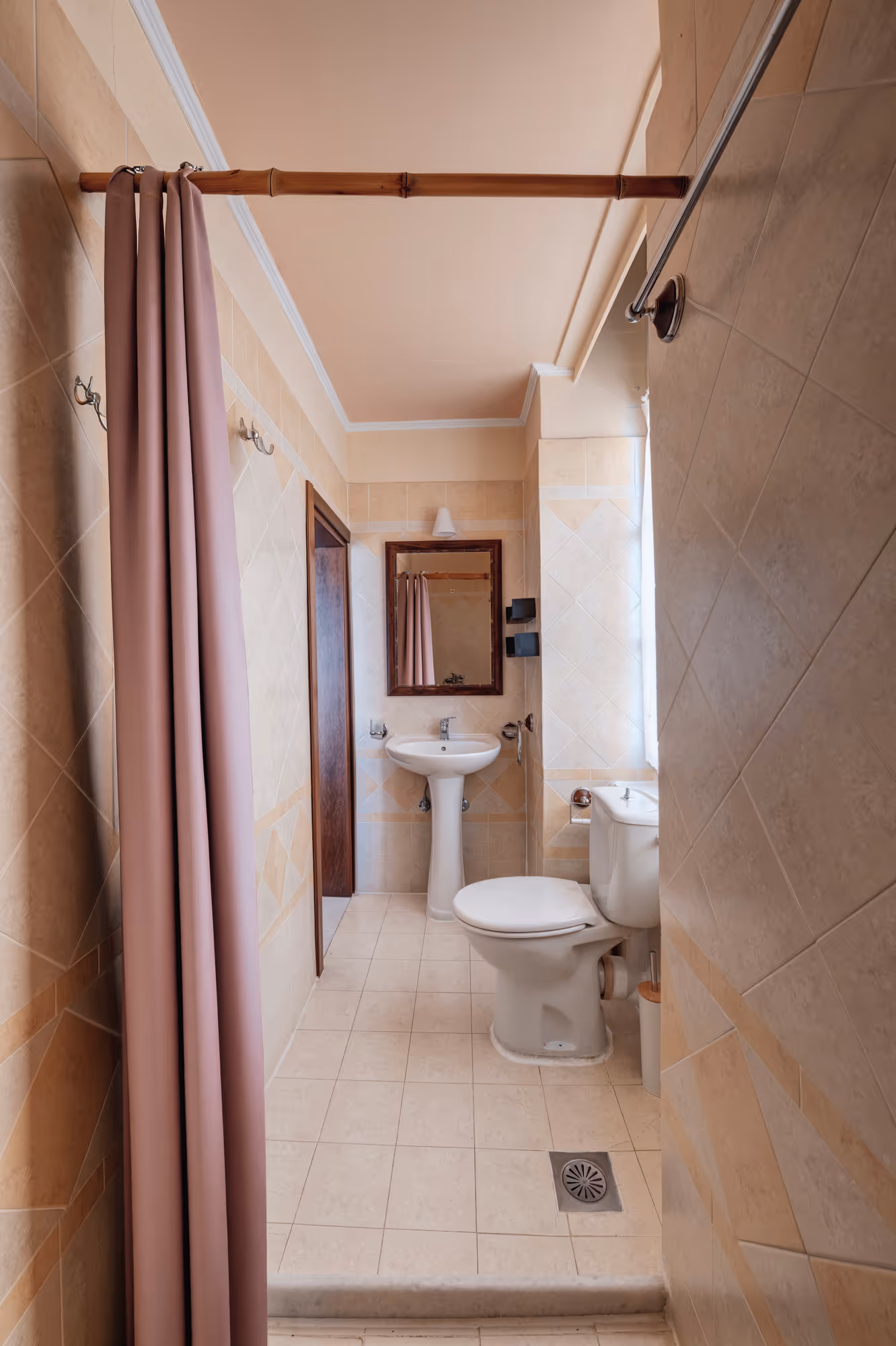 Small bathroom with beige tiles, a white toilet, pedestal sink, mirror, and a purple shower curtain on a bamboo rod.