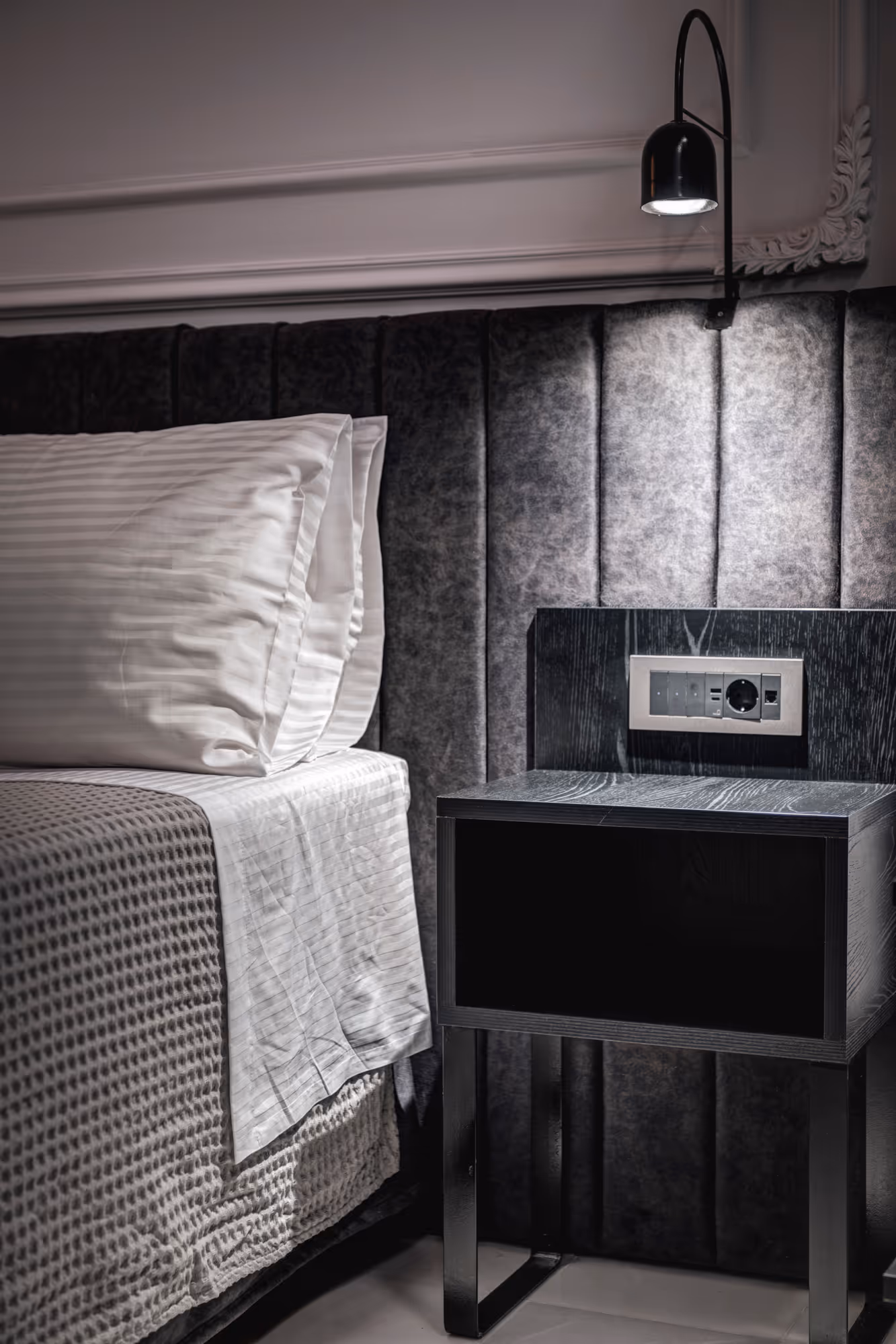 Modern bedroom corner with white striped pillows, textured bedding, black nightstand, and a wall-mounted lamp with power outlets.