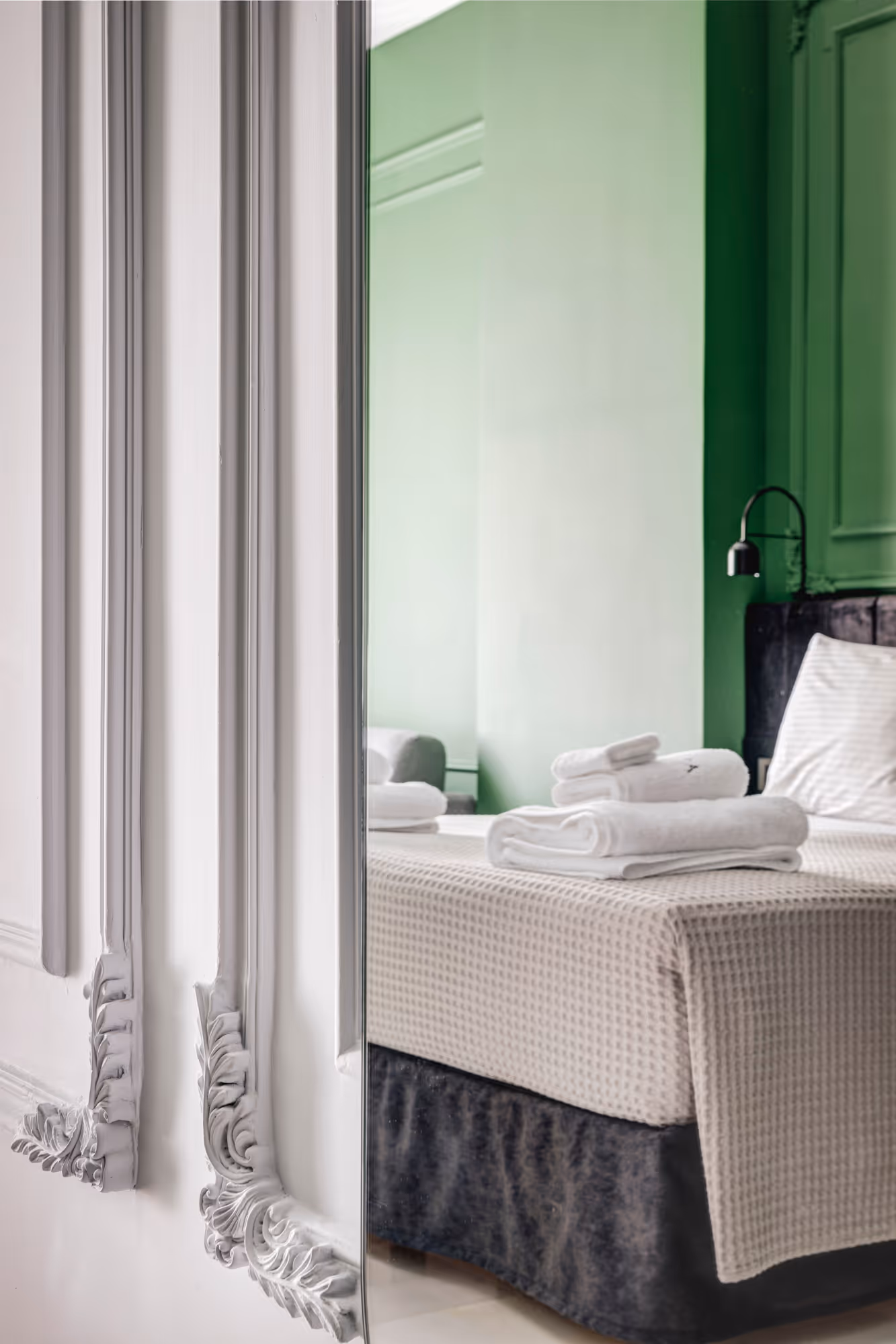 Reflection of a bedroom with a bed covered in beige textured bedding, a pillow, folded white towels, and green walls as seen in an ornate mirror.