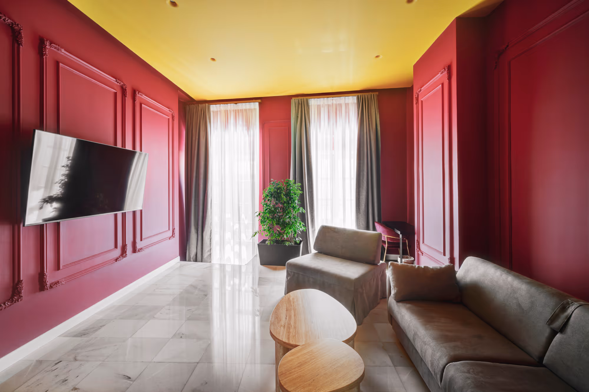 Modern living room with rich burgundy walls, bright yellow ceiling, gray sofa and chairs, sheer curtains, and two wooden tables.