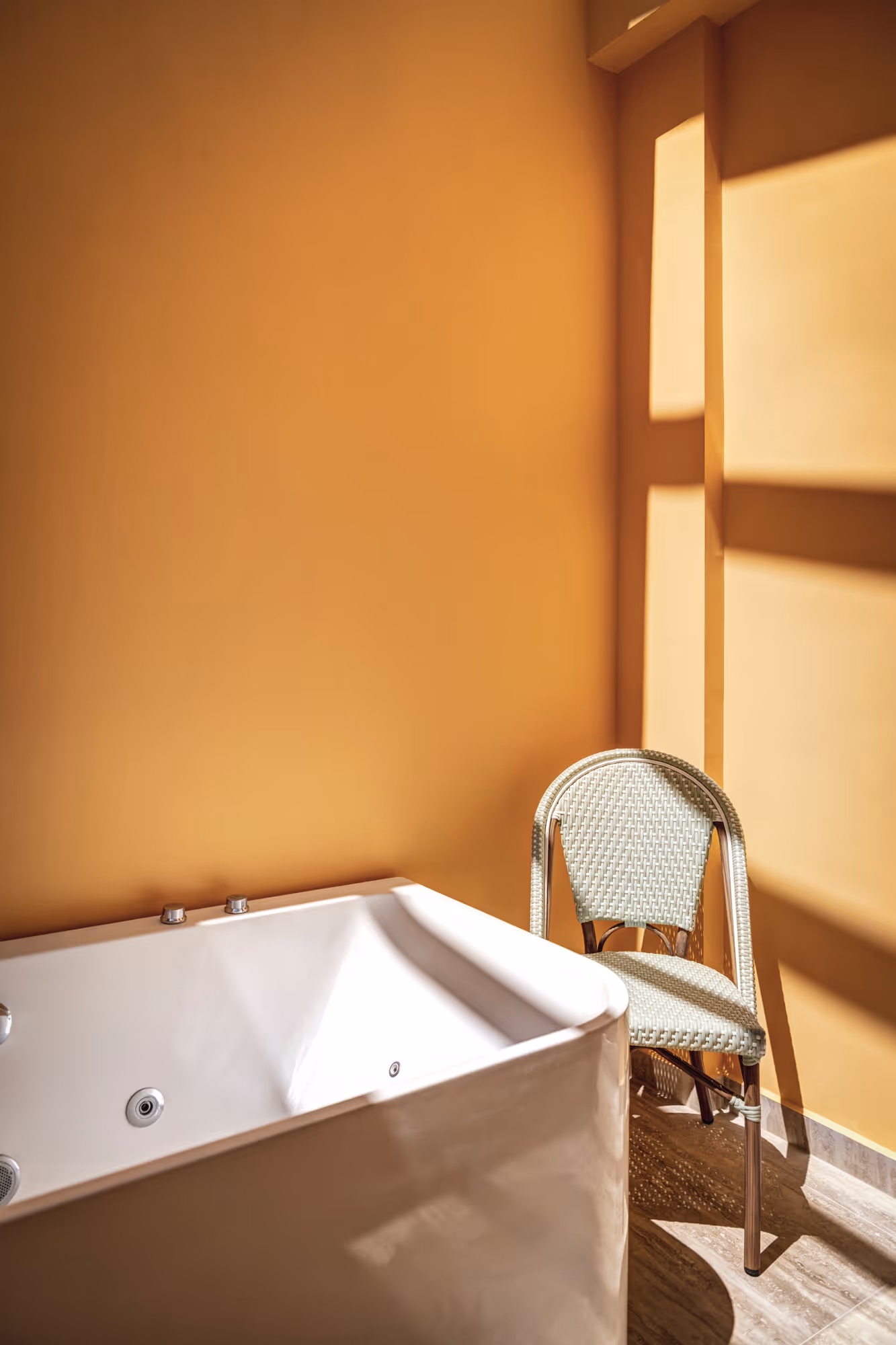 White bathtub and a woven chair beside mustard yellow walls with sunlight casting window shadows.