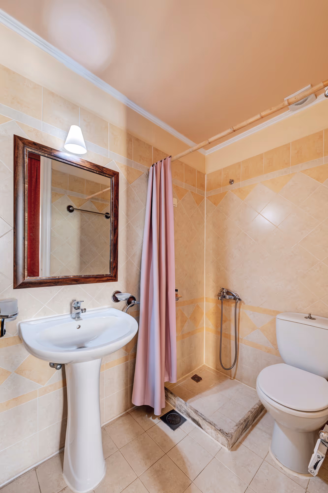 Small bathroom with beige tiled walls, white pedestal sink, wooden framed mirror, pink shower curtain, and white toilet.