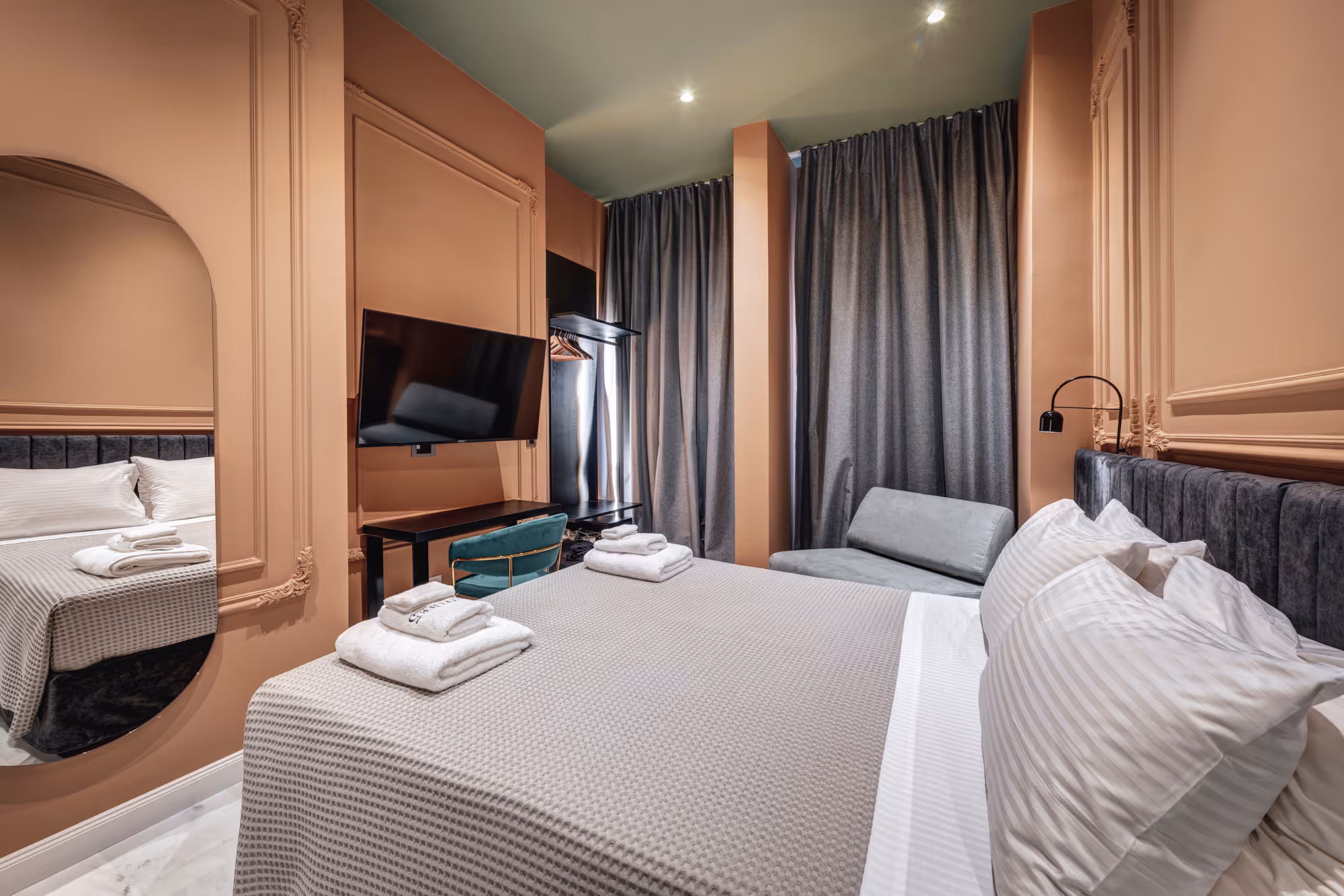 Modern hotel room with beige walls, a large bed with a gray blanket and white pillows, a wall-mounted TV, a teal chair, and a gray sofa near dark curtains.