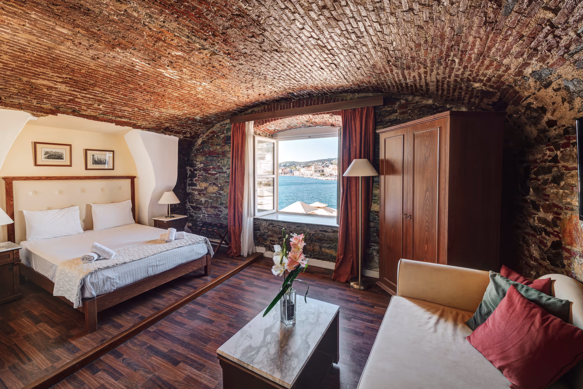 Cozy bedroom with arched brick ceiling, a bed with white linens, a window overlooking water, and a small sitting area with sofa and pillows.