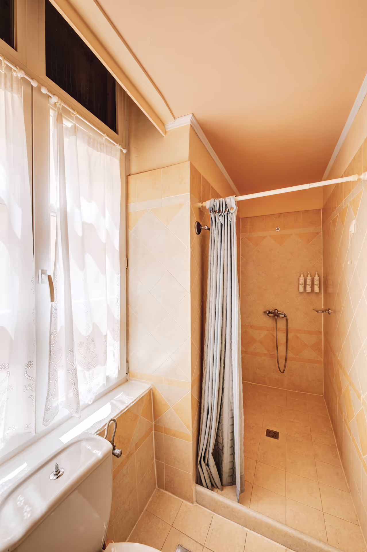 Small beige tiled bathroom with a white toilet by a window with sheer curtains and a shower area with a striped curtain and mounted shower fixtures.