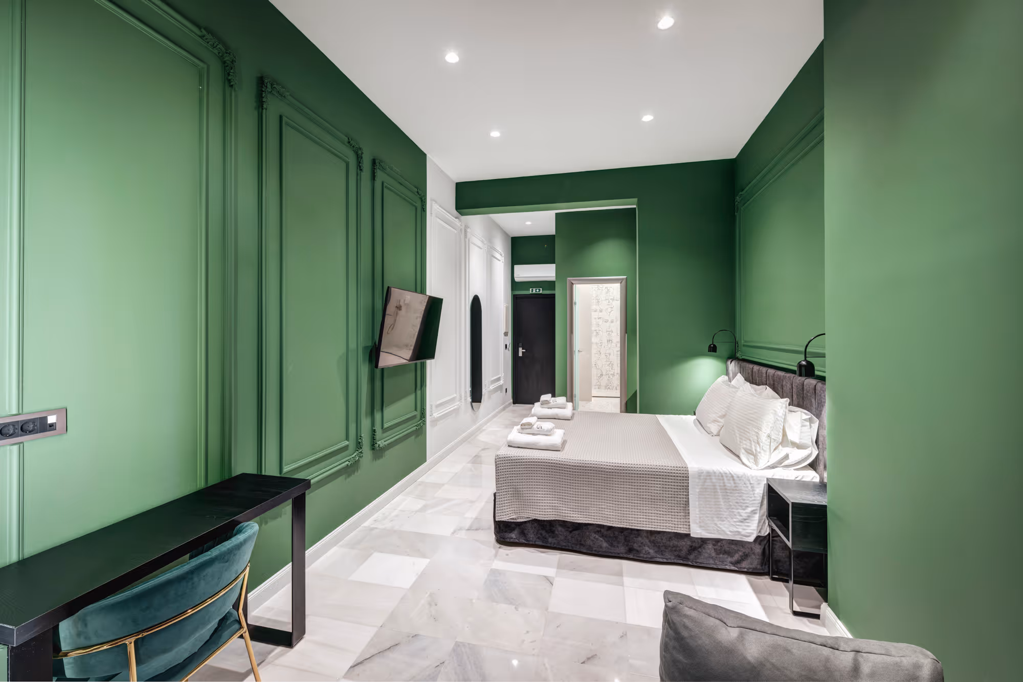 Modern bedroom with green walls, a gray bed with white and gray bedding, a mounted TV, and a black desk with a green chair.