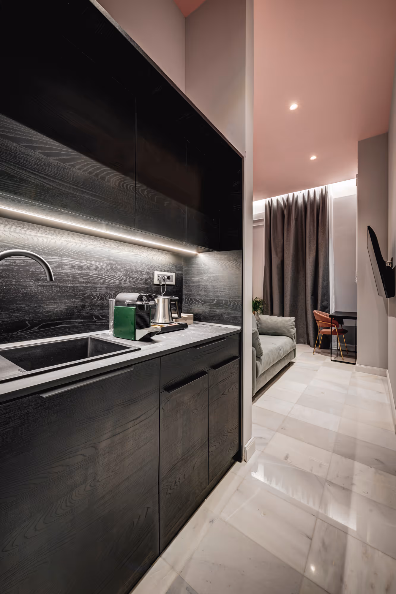 Modern kitchenette with dark wood cabinets, built-in sink, coffee machine, and marble floor leading to a living area with a gray sofa and a workspace with a red chair and dark curtains.