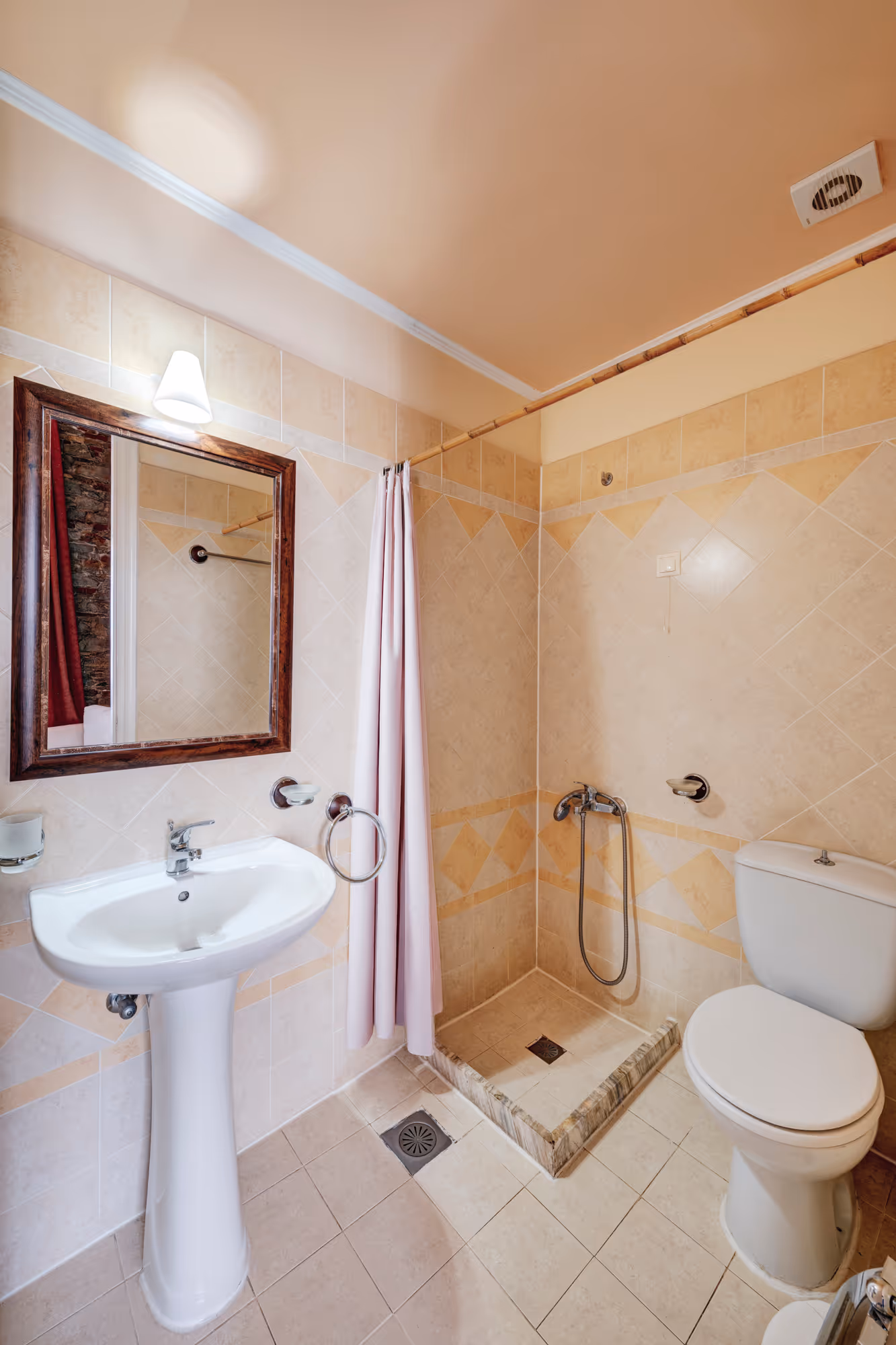 Small beige tiled bathroom with a pedestal sink, mirror, shower area with curtain, and toilet.
