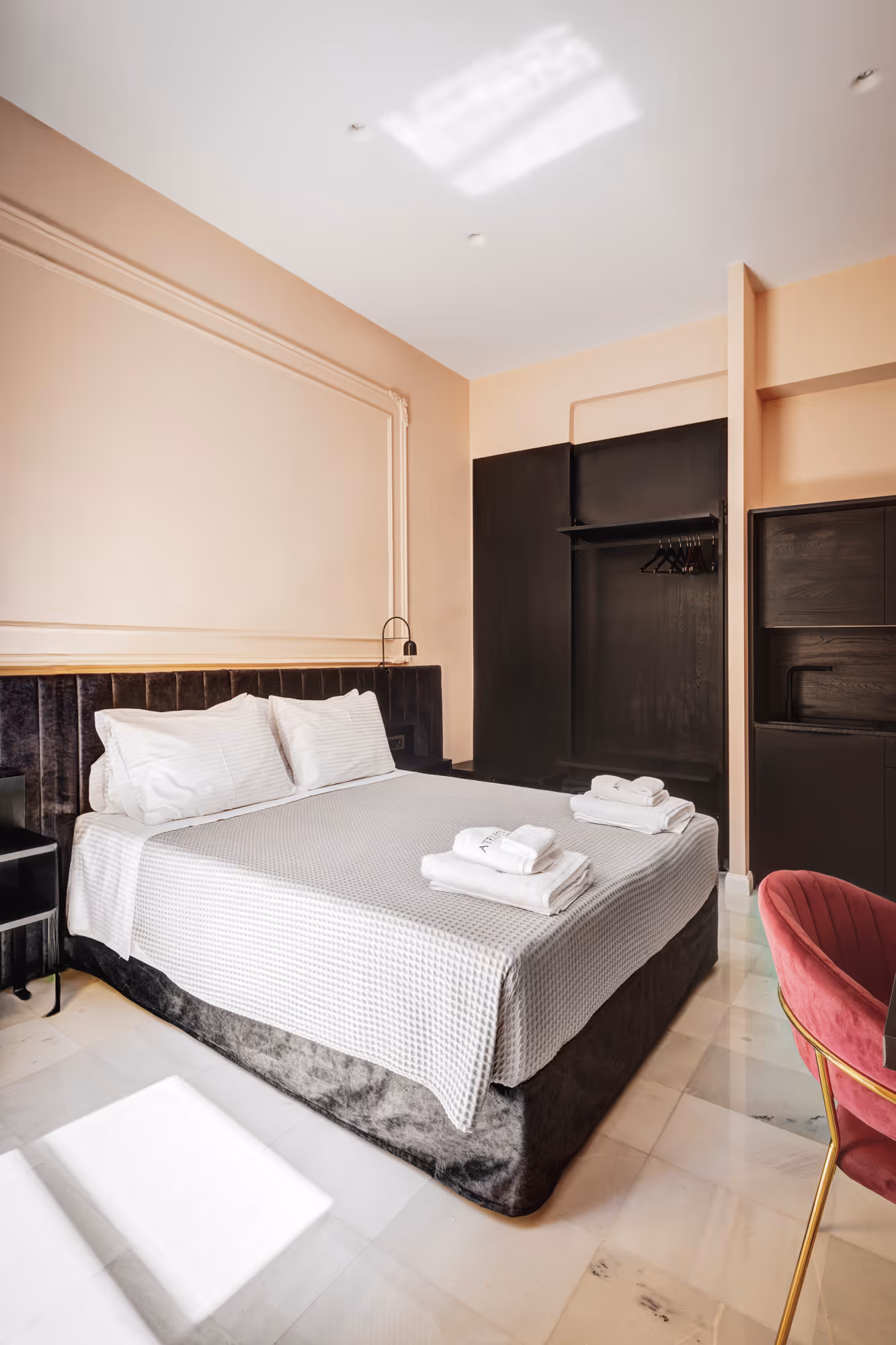 Minimalist hotel room with a neatly made bed covered in a gray textured blanket, white pillows, folded towels on top, black closet, and a red velvet chair.