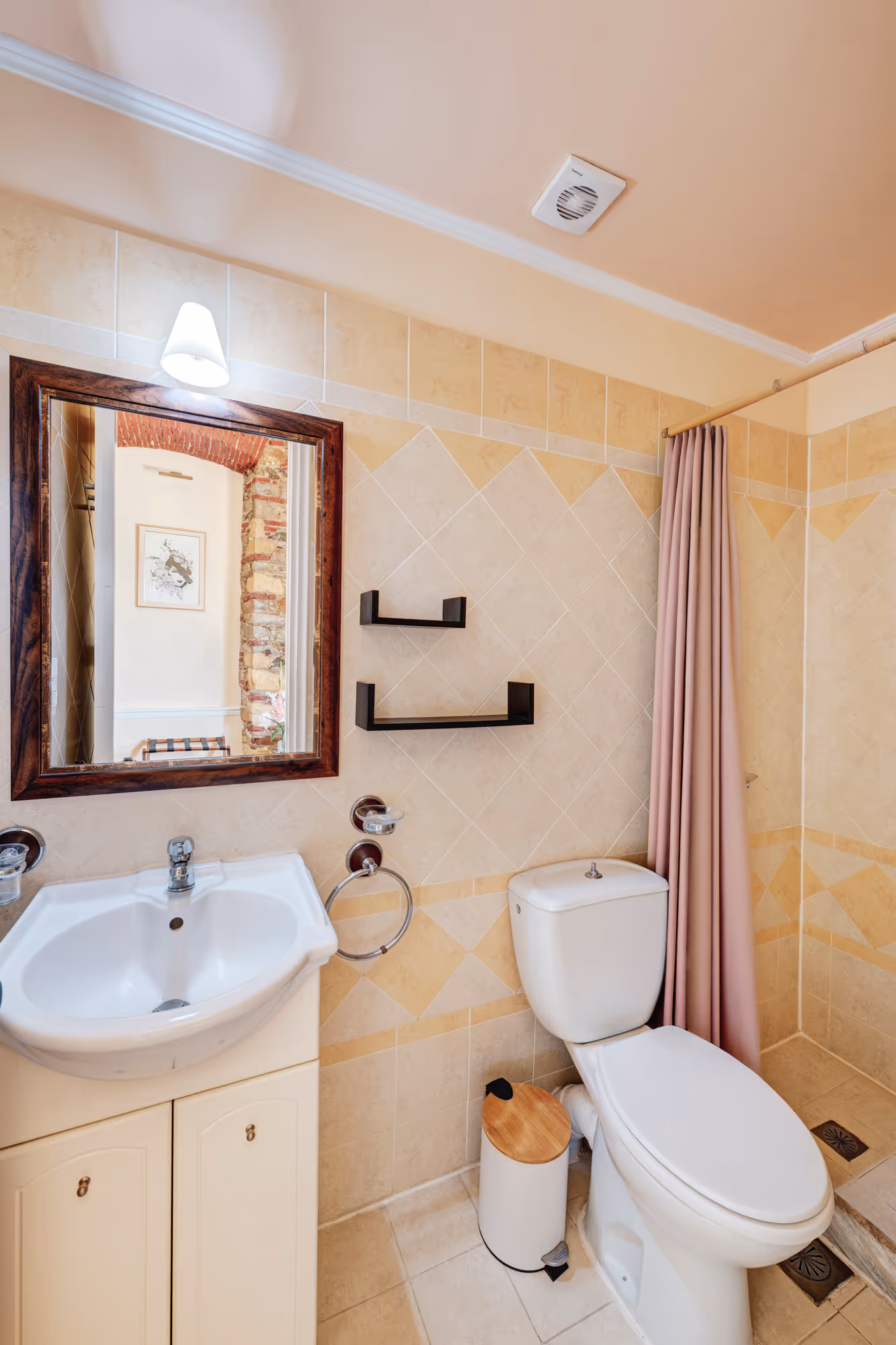 Bathroom with white sink and cabinet, framed mirror, white toilet, wooden-lid trash bin, and beige tiled walls and floor.