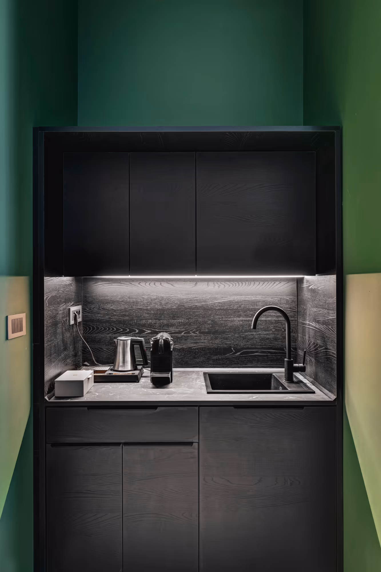 Compact modern kitchenette with black cabinetry, marble countertop, sink with black faucet, electric kettle, and coffee machine with under-cabinet lighting.