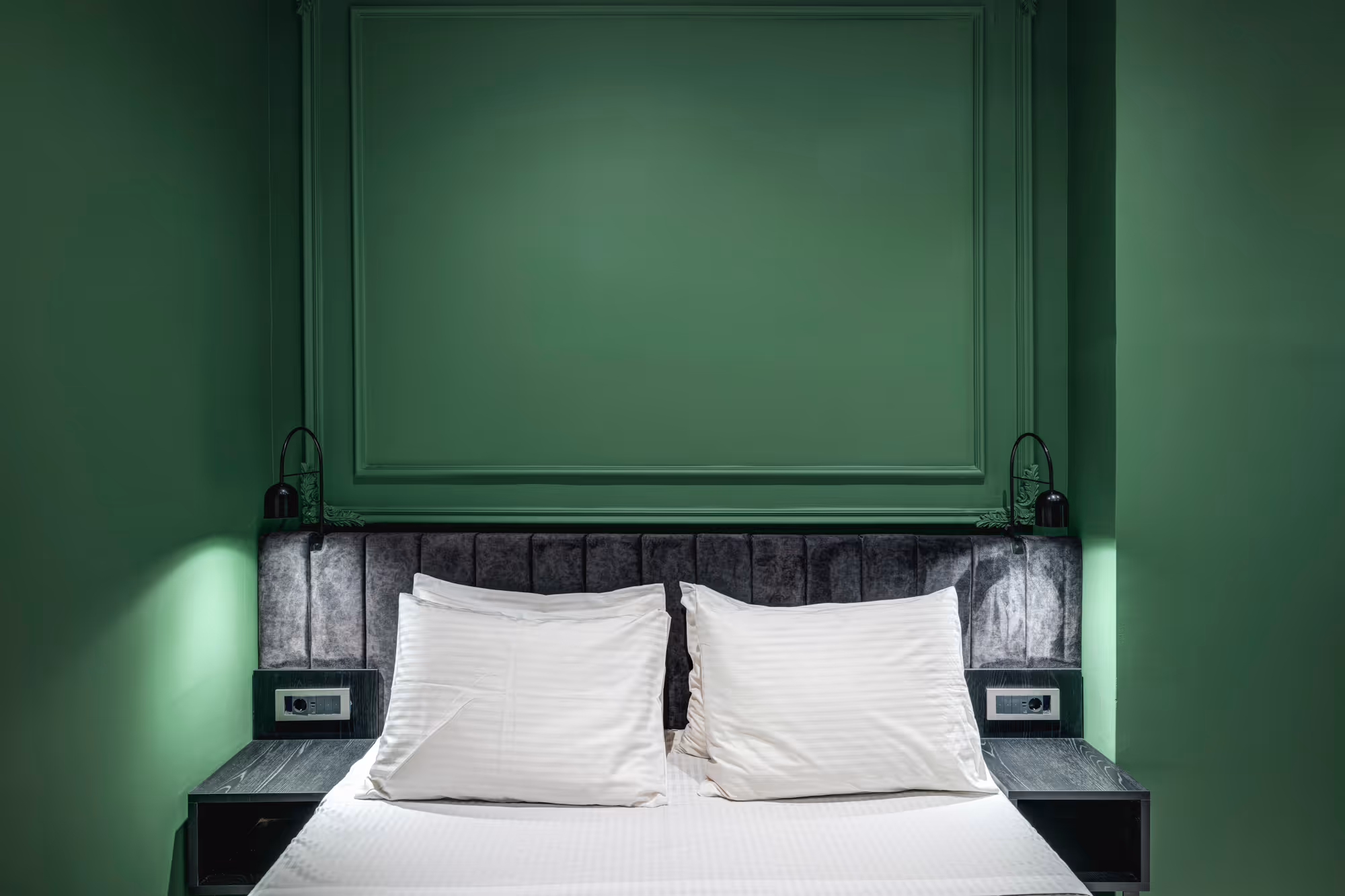 Modern bed with white pillows and linens against a dark gray upholstered headboard and green paneled wall.
