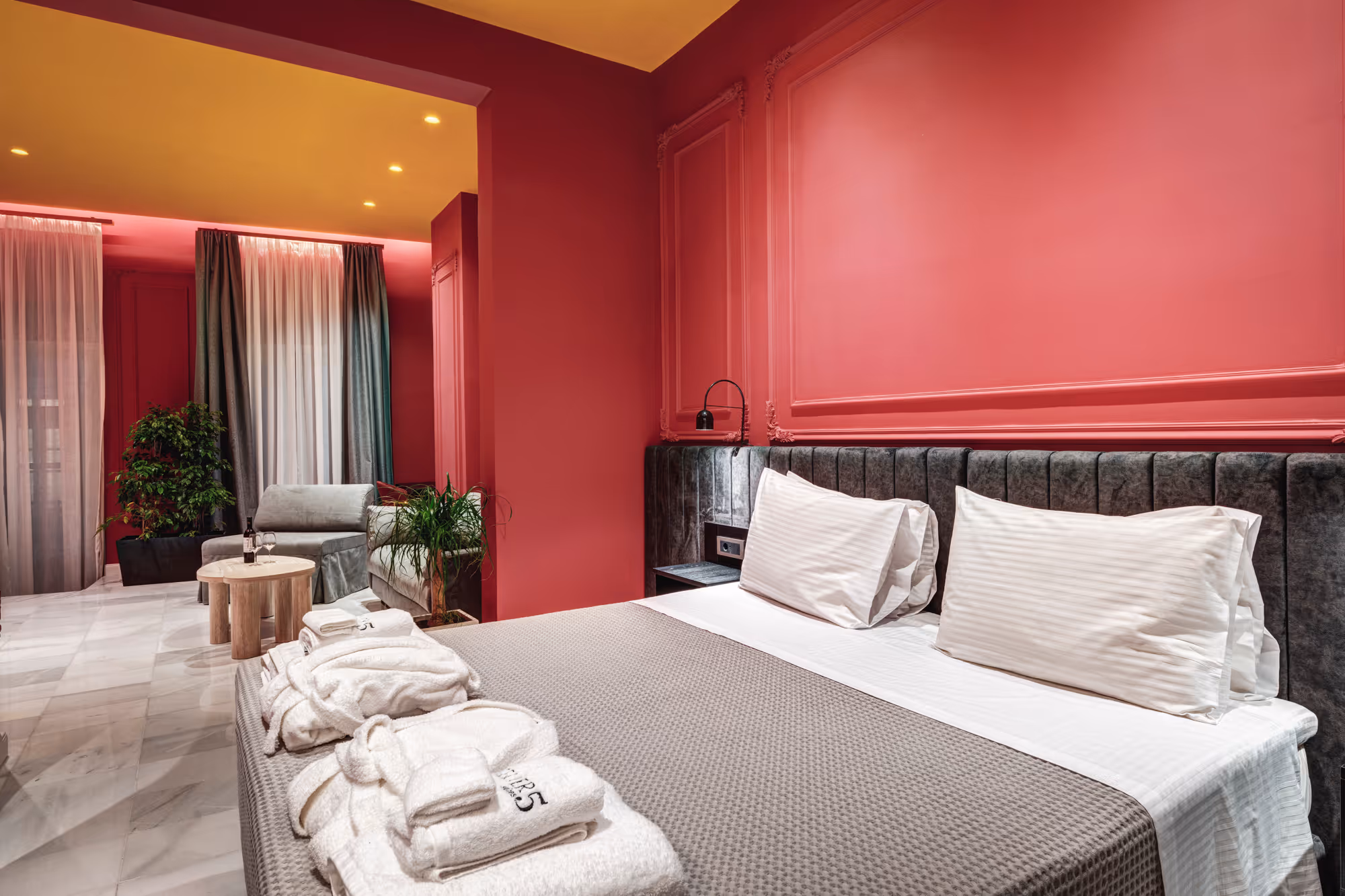 Modern bedroom with large bed, gray headboard, white pillows, robes, and pink walls with plants and chairs.